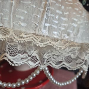May include: A close-up of a vintage lampshade featuring cream-coloured fabric with white accents and a delicate lace trim. A strand of faux pearls hangs from the bottom. The lampshade sits on a red and white polka-dotted surface, adding a touch of elegance.