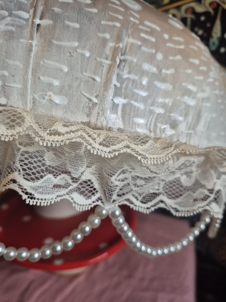 May include: A close-up of a vintage lampshade featuring cream-coloured fabric with white accents and a delicate lace trim. A strand of faux pearls hangs from the bottom. The lampshade sits on a red and white polka-dotted surface, adding a touch of elegance.