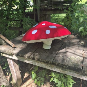 Peut inclure: Un jouet champignon en peluche doté d'un large chapeau rouge arrondi orné de taches circulaires blanches. Le dessous du chapeau est de couleur crème claire, et le pied est d'une couleur crème assortie, formant une base solide pour la structure.