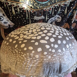 May include: A large, mushroom-shaped decorative cushion with a grey top covered in white spots. The cushion has a white lace trim around the bottom edge. The cushion is resting on a pink surface.