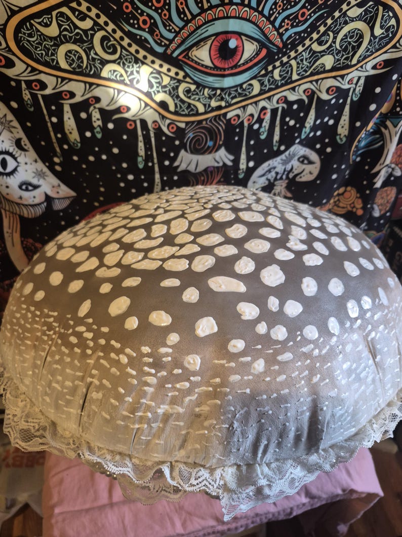 May include: A large, mushroom-shaped decorative cushion with a grey top covered in white spots. The cushion has a white lace trim around the bottom edge. The cushion is resting on a pink surface.