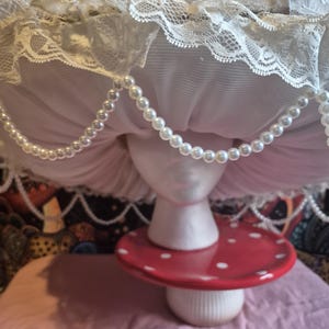 May include: A decorative mushroom ornament. The mushroom features a red cap with white spots, a white stem, and a white, lace-trimmed top. A string of faux pearls hangs from the top. It is displayed on a pink surface.