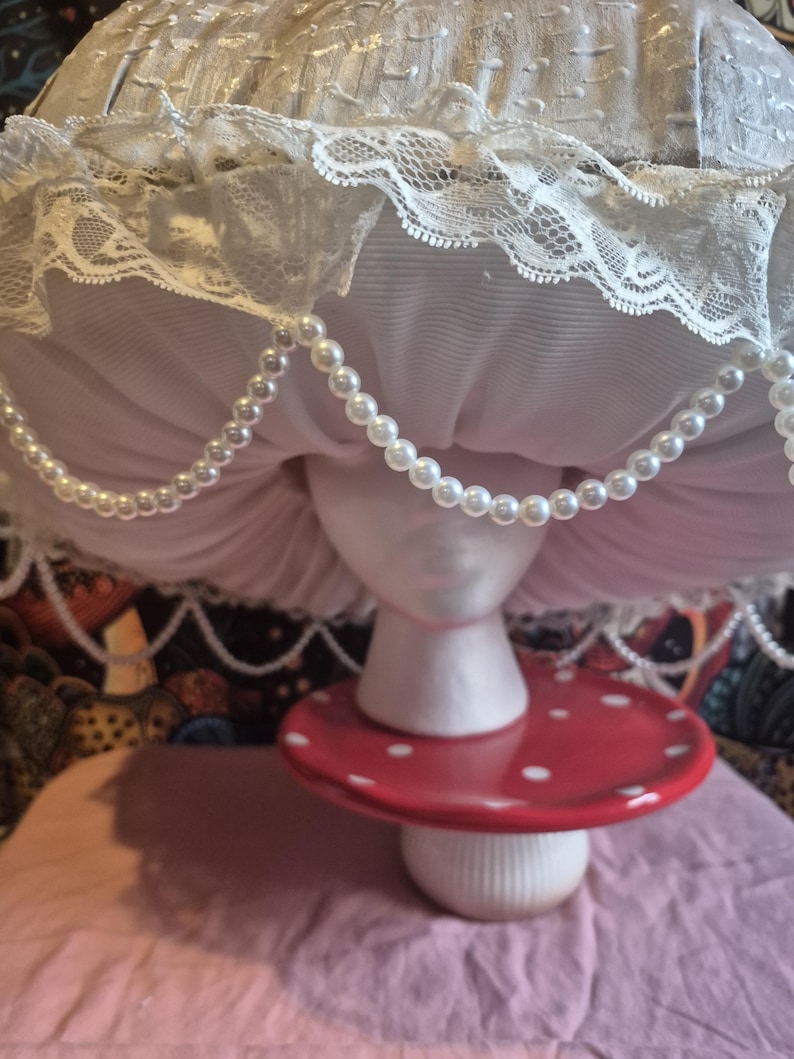 May include: A decorative mushroom ornament. The mushroom features a red cap with white spots, a white stem, and a white, lace-trimmed top. A string of faux pearls hangs from the top. It is displayed on a pink surface.