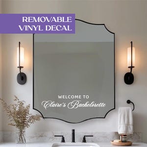 May include: A white vinyl decal applied to a scalloped-edge mirror, featuring the text "WELCOME TO Claire's Bachelorette" in a combination of sans-serif and script fonts. The decal is positioned centrally on the reflective glass surface.