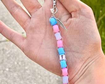 May include: A beaded keychain with a silver clasp and a feather charm. The beads are in shades of pink, blue, and white, with a white letter "T" bead. The keychain is held up against a blurred green background.