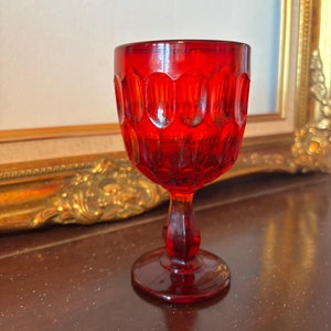 May include: A red glass goblet with a stemmed base and a textured design. The goblet is sitting on a dark wooden surface.