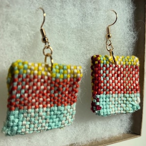 May include: Handmade woven earrings with gold-colored hooks. The earrings feature a square shape with a colorful woven pattern. The colors include red, yellow, blue, and white. The earrings are displayed in a small, white-lined box.