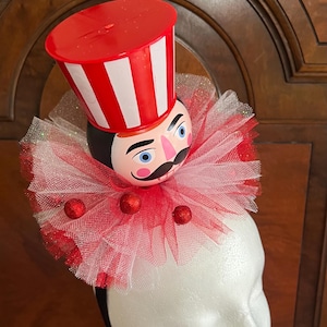 May include: A headband featuring a nutcracker head with a black mustache and blue eyes, topped with a red and white striped plastic hat. A large, layered red and white tulle ruff collar surrounds the neck, accented with small red glittery spheres.