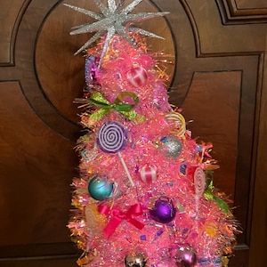 May include: A pink tinsel cone-shaped decoration topped with a silver glitter star. The surface is adorned with various colourful ornaments, including round baubles, small bows, and lollipop-shaped accents in shades of purple, blue, and pink.