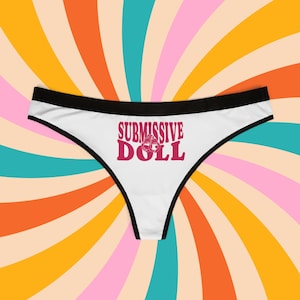 Könnte beinhalten: Weiße Spitzen-String-Unterwäsche mit schwarzem Bund und dem aufgedruckten Text "SUBMISSIVE DOLL" in Pink.