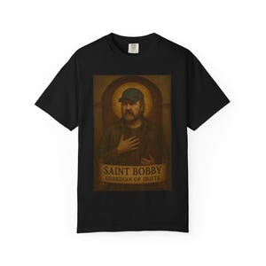 May include: Black t-shirt featuring a portrait of a man wearing a baseball cap, with the text "SAINT BOBBY GUARDIAN OF IDJITS" on a banner below the portrait. The portrait has a religious art style.