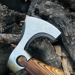 May include: A silver axe head with intricate engraved designs, including a circular emblem and Viking-style patterns. The axe features a dark gray blade and a wooden handle with a rich, brown grain. The axe is displayed against a backdrop of weathered wood.