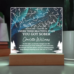 Puede incluir: Luz de noche personalizada con base de madera. El panel acrílico transparente presenta un diseño de cielo estrellado con el texto "UNDER THESE BEAUTIFUL STARS YOU GOT SOBER" y un nombre. Incluye la fecha y el lugar.