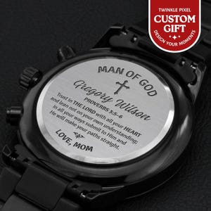 May include: Close-up of a black wristwatch with a silver-colored face. The watch face is engraved with the words "MAN OF GOD", a cross, the name "Gregory Wilson", and a Bible verse. The watch also has the words "LOVE, MOM".