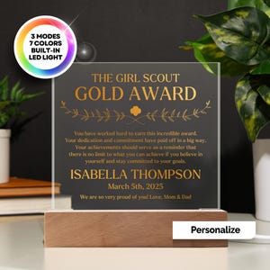 May include: A personalized Girl Scout Gold Award plaque with a wooden base and LED lighting. The plaque features gold text that reads "The Girl Scout Gold Award" and includes a message, recipient's name, and date. The plaque has 3 modes and 7 colors.