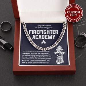 Puede incluir: Un collar de cadena plateada presentado en una caja de regalo roja. La caja contiene una tarjeta azul marino con el texto "Felicitaciones por completar tu FIREFIGHTER ACADEMY" y un gráfico de boca de incendios. También se ven un anillo y un bolígrafo.