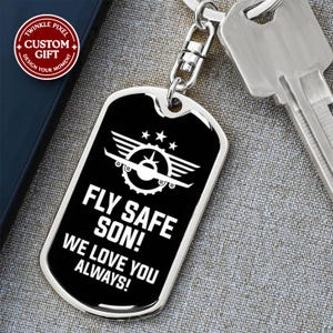 May include: A silver-toned metal dog tag keyring featuring a black rectangular insert with rounded corners. The design includes a white graphic of an aeroplane with wings and three stars above it, followed by the text FLY SAFE SON! WE LOVE YOU ALWAYS!
