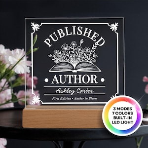 May include: A clear acrylic plaque with the words "PUBLISHED AUTHOR" and the name "Ashley Carter" engraved in white. The plaque features a floral design and sits on a wooden base. The LED light has 3 modes and 7 colors.
