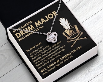 May include: A silver necklace with a sparkling pendant in a gift box. The box is black with white text that reads "TO AN AMAZING DRUM MAJOR WHO INSPIRED US ALL." The box also has a graphic of a drum and drumsticks.