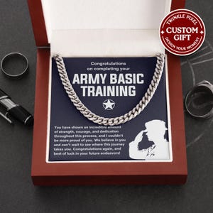 May include: A silver chain necklace displayed in a red gift box. The box contains a card with the text "Congratulations on completing your ARMY BASIC TRAINING" and a silhouette of a soldier saluting. A ring and pen are also visible.