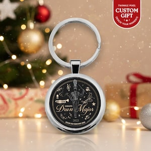 May include: A silver keychain with a round pendant featuring a black and gold design. The pendant has a drum major illustration and the text "Drun Major 2025". The keychain is a custom gift with the text "Twinkle Pixel Custom Gift".