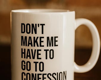 May include: White ceramic mug with the text "DON'T MAKE ME HAVE TO GO TO CONFESSION" in black. The mug has a curved handle and is set against a blurred background.
