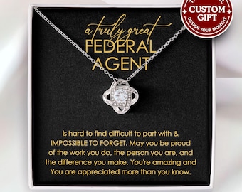 May include: A silver-colored necklace with a pendant in a gift box. The pendant has a central clear stone surrounded by smaller stones. The box has text that reads "a truly great FEDERAL AGENT". The gift box also has a message.