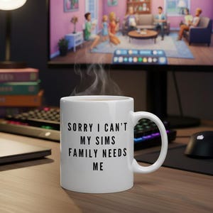 May include: A white ceramic mug with the text "SORRY I CAN'T MY SIMS FAMILY NEEDS ME" in black. Steam rises from the mug, suggesting a hot beverage. The mug sits on a wooden desk with a computer, keyboard, and mouse in the background.
