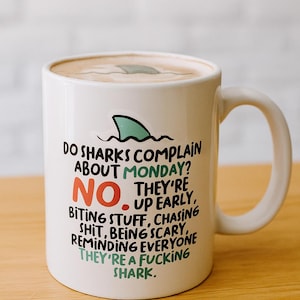 May include: White ceramic mug with a handle, featuring the text "DO SHARKS COMPLAIN ABOUT MONDAY? NO. THEY'RE UP EARLY, BITING STUFF, CHASING SHIT, BEING SCARY, REMINDING EVERYONE THEY'RE A FUCKING SHARK." and a shark fin graphic.