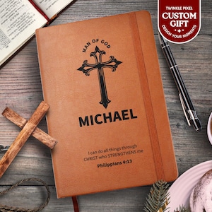 Puede incluir: Un diario de cuero marrón personalizado con el nombre "MICHAEL" y un diseño de cruz negra. El diario incluye el texto "Man of God" y un versículo bíblico. También se ven una cruz de madera y un bolígrafo.