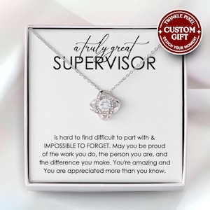May include: A silver necklace with a knot pendant, presented in a gift box. The box has text that reads "a truly great SUPERVISOR" and a message below. A circular sticker says "CUSTOM GIFT". The pendant has a central clear stone.