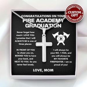 Può includere: Una collana con pendente a croce argentata esposta in una confezione regalo. La scatola presenta il testo "Congratulazioni per la tua laurea all'Accademia dei Vigili del Fuoco" e un messaggio da "Mamma".