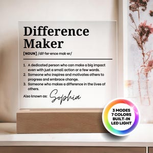 Puede incluir: Una placa de acrílico transparente con texto negro que define el término Difference Maker, incluyendo el nombre Sophia en cursiva. La placa se asienta sobre una base de madera rectangular e incluye un gráfico circular 3 MODES 7 COLORS.