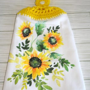 May include: White hanging kitchen towel with a watercolor sunflower design. The towel features yellow crochet detailing at the top and a button closure. The sunflowers are yellow and orange with green leaves.