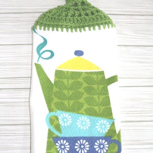 May include: A white hanging kitchen towel with a green crocheted top featuring a white button. The towel displays a graphic print of a green teapot with a yellow lid and two stacked teacups in light blue and dark blue, each with white floral patterns.