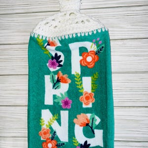 May include: A teal kitchen towel with the word "SPRING" in white block letters, decorated with orange, pink, and purple floral accents. The top portion is white crochet with a button closure.