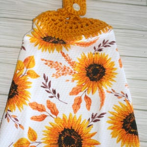 May include: White kitchen towel with a sunflower and autumn leaf pattern. The towel has a crocheted mustard yellow top with a button closure for hanging. The sunflowers are yellow and orange with dark brown centers.