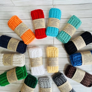 May include: A collection of rectangular crochet cast iron skillet handle covers in various colors including orange, red, light blue, teal, black, white, yellow, green, dark green, gray, brown, and navy blue, each wrapped with a paper label.