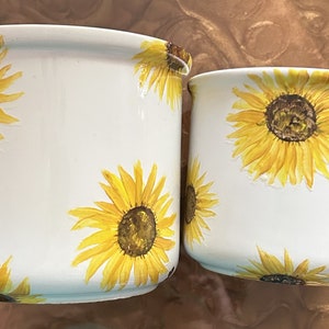 May include: Two white ceramic planters with a sunflower design. The larger planter has a diameter of approximately 6 inches and the smaller planter has a diameter of approximately 4 inches.