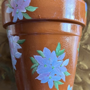 May include: A terracotta flower pot with a hand-painted design of blue and purple flowers. The pot is decorated with green leaves and has a glossy finish.
