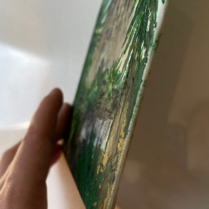 May include: A small, rectangular painting on canvas. The artwork depicts a landscape scene with green foliage, a light-colored path, and a blue sky. The edges of the canvas are visible, and the painting is held at an angle.