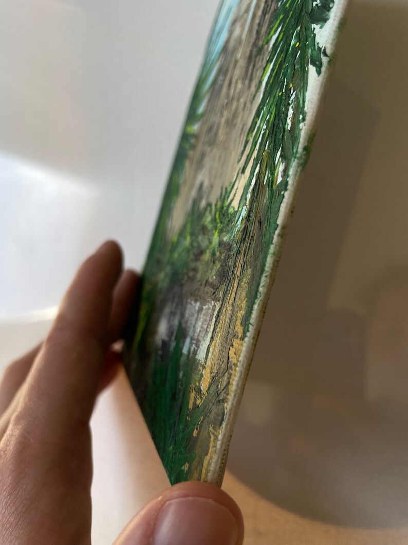 May include: A small, rectangular painting on canvas. The artwork depicts a landscape scene with green foliage, a light-colored path, and a blue sky. The edges of the canvas are visible, and the painting is held at an angle.