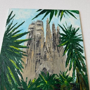 May include: An oil painting depicts a towering cathedral with multiple spires, rendered in shades of gray and beige. Lush green palm fronds frame the building against a light blue sky. The artwork showcases a textured, impressionistic style.