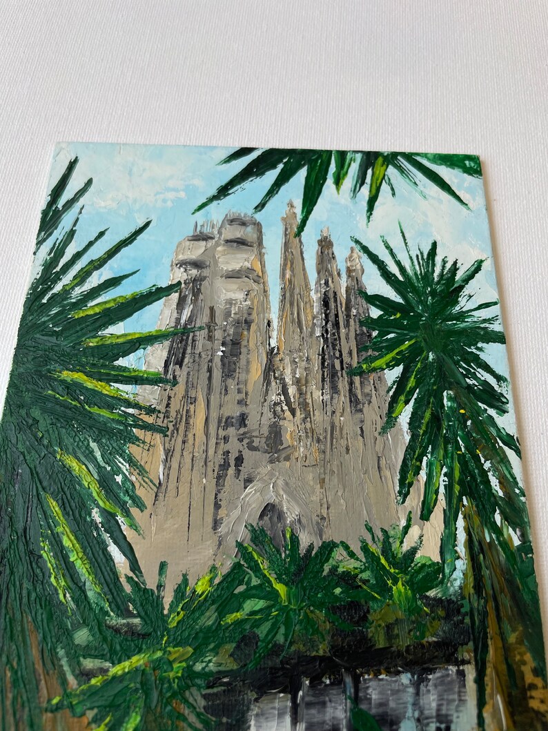 May include: An oil painting depicts a towering cathedral with multiple spires, rendered in shades of gray and beige. Lush green palm fronds frame the building against a light blue sky. The artwork showcases a textured, impressionistic style.