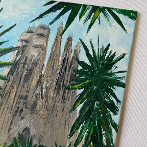 May include: An oil painting depicts the Sagrada Familia basilica in Barcelona, Spain, viewed from a low angle. The artwork features the church's gray stone spires against a light blue sky, framed by green palm fronds. The painting has a textured, impasto style.