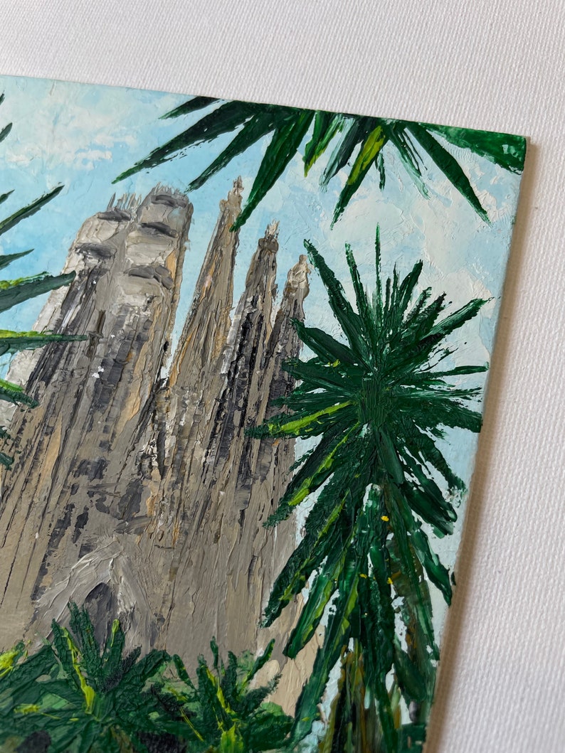 May include: An oil painting depicts the Sagrada Familia basilica in Barcelona, Spain, viewed from a low angle. The artwork features the church's gray stone spires against a light blue sky, framed by green palm fronds. The painting has a textured, impasto style.