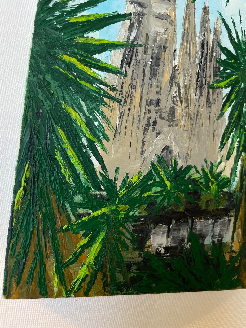 May include: An oil painting depicts the Sagrada Familia basilica in Barcelona, Spain. The artwork features the church's spires and facade, framed by green palm fronds. The sky is a light blue, and the painting has a textured, impressionistic style.
