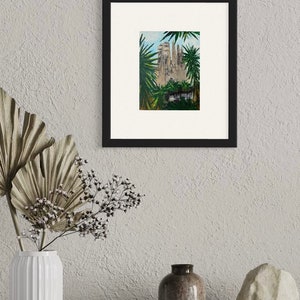 May include: A framed artwork featuring a painting of a cathedral, framed in black. The painting depicts a detailed architectural structure surrounded by green foliage. The artwork is displayed on a neutral-toned wall, with decorative elements in the foreground.