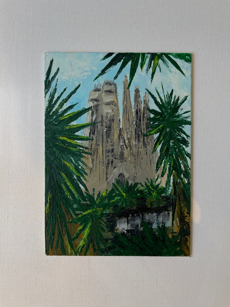 May include: An oil painting of the Sagrada Familia basilica in Barcelona, Spain. The artwork depicts the cathedral's intricate facade and spires, framed by lush green palm fronds against a light blue sky. The painting is on a white canvas.
