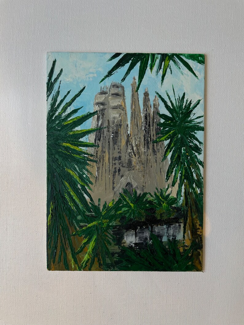 May include: An oil painting of the Sagrada Familia in Barcelona, Spain, framed by green palm fronds. The cathedral is depicted in shades of gray and beige against a light blue sky. The artwork is on a white canvas.